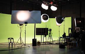 Behind the scenes of a green screen set with a video camera, monitor, and a set of many types of lighting equipments on tripods, set up for shooting video